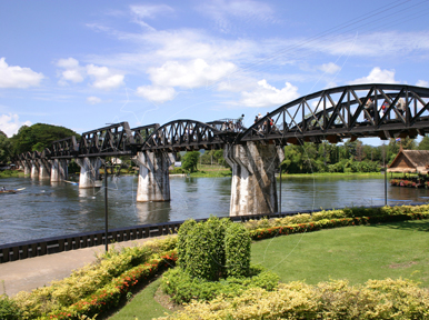 Black steel truss railway bridge over the River Kwai in Kanchanaburi, Thailand, with landscaped garden in foreground