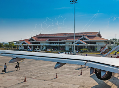 Passengers walk across sunlit tarmac past an aircraft wing toward a small red-roofed regional airport terminal.