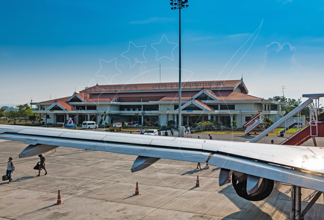 Aircraft wing over tarmac at a small Thai regional airport terminal with red tiled roof, passengers walking to steps