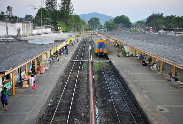 Elevated view of a rural railway station with covered platforms, twin tracks and a blue-yellow diesel railcar, hills beyond