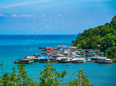 Stilt-house fishing village built over turquoise sea, viewed from a forested headland under a clear blue sky