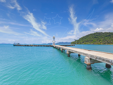Concrete pier stretching over turquoise sea toward a small lighthouse, with forested island and moored boat