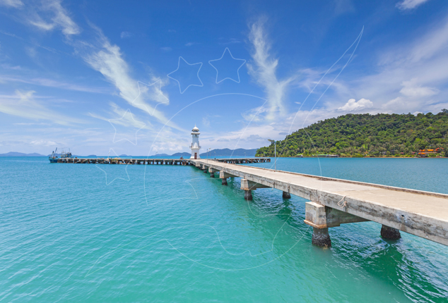 Long concrete pier with small white lighthouse stretching over turquoise sea toward a green forested island