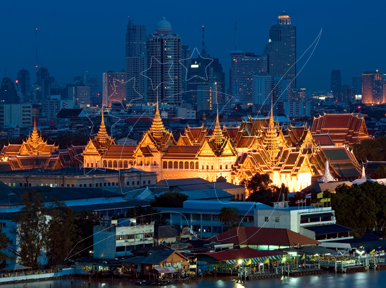Bangkok's Grand Palace illuminated at dusk with city skyscrapers behind and the Chao Phraya River in front