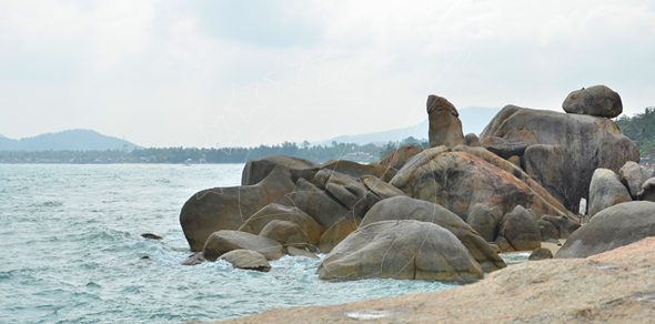 Weathered granite boulders on a rocky shoreline with turquoise sea, distant hills and hazy sky