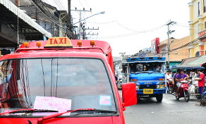 Red songthaew truck with TAXI roof sign parked on a busy Thai street, blue songthaew and motorbikes behind.