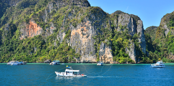White wooden fishing boat crossing turquoise sea below jungle-covered limestone karst cliffs, boats moored near shore