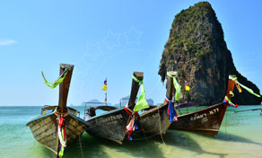 Traditional wooden longtail boats moored on white sand beach below a towering limestone karst cliff, Thailand