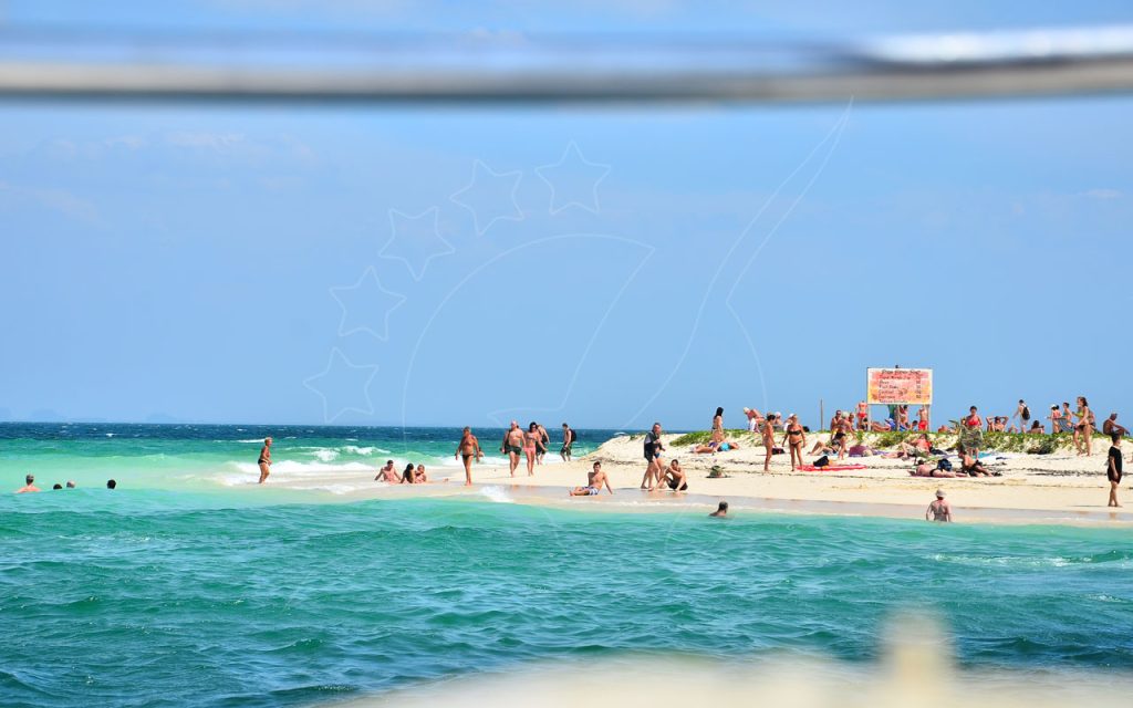 Tourists sunbathing and swimming on a narrow tropical sandbar with turquoise sea and a drinks price sign