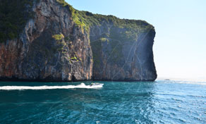 Speedboat crossing turquoise sea beside a towering limestone cliff island covered in green vegetation