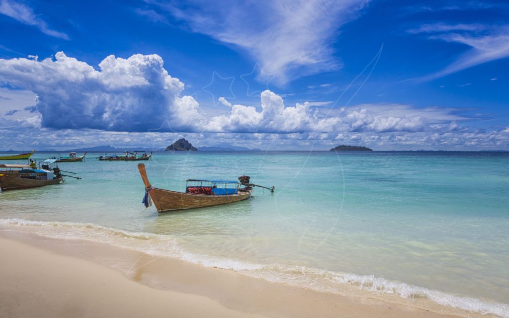 Wooden longtail boats moored in clear turquoise shallows off a white sand beach, islands on the horizon
