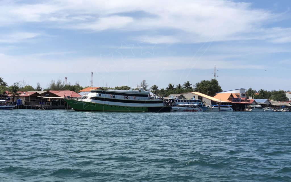 Green and white passenger ferry moored at a pier lined with stilt buildings, viewed across choppy sea