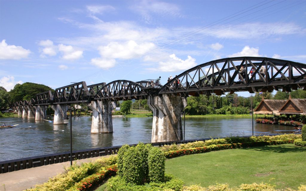 Black steel truss Bridge over the River Kwai in Kanchanaburi, Thailand, with tourists walking across under blue sky