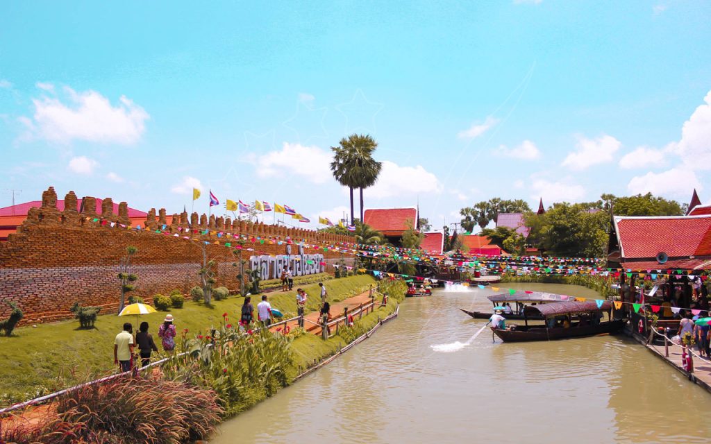 Long-tail boats on a canal beside a brick wall and colourful bunting at a Thai floating market, tourists walking a path