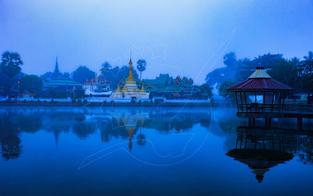 Golden Thai temple and pavilion reflected in a misty lake at blue hour, Mae Hong Son-style lakeside town scene