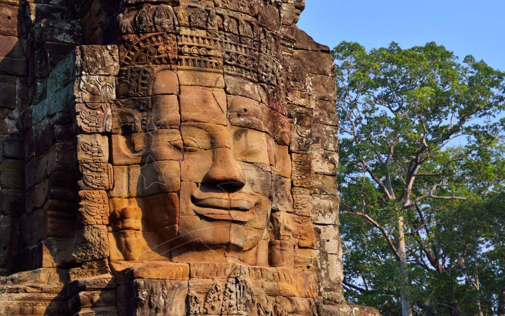 Giant carved sandstone face on a Bayon temple tower at Angkor Thom, Cambodia, with green tree and blue sky