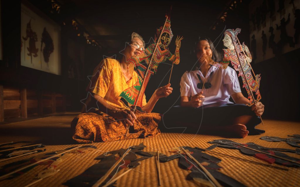Elderly woman and young student hold ornate Thai shadow puppets on a woven mat in a dim wooden hall