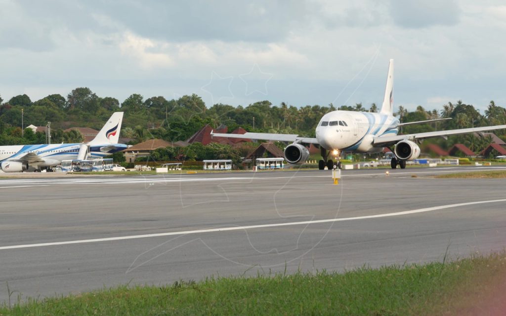 Bangkok Airways jet taxiing on tropical airport runway with palm trees and thatched-roof terminal buildings