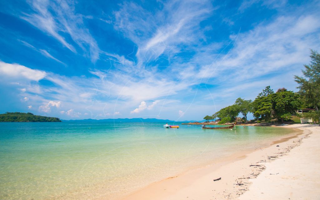 White sand beach with clear turquoise shallows, moored longtail boat and speedboat, tree-lined headland under blue sky