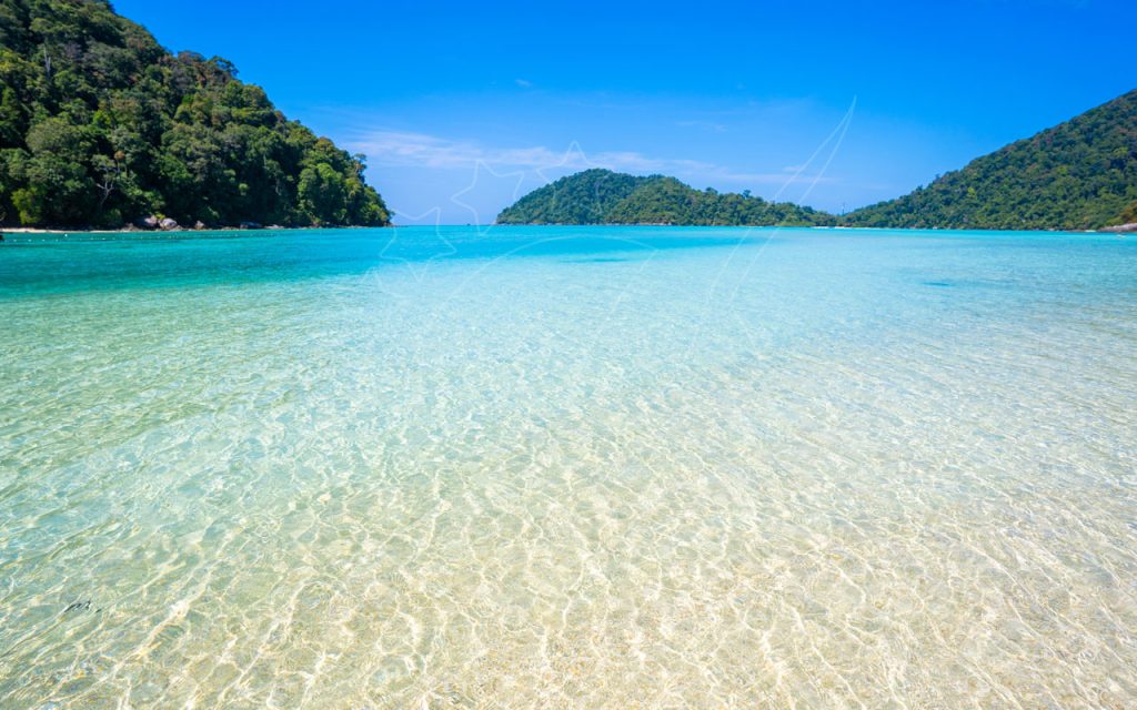 Crystal-clear turquoise shallows over pale sand with jungle-covered islands and blue sky, tropical bay