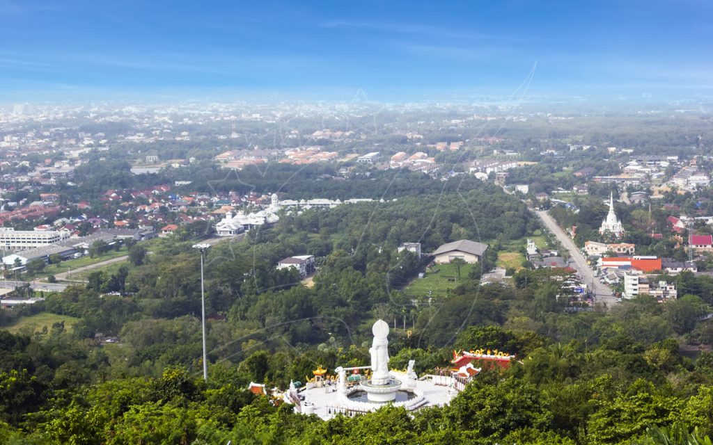 Hilltop white Guanyin statue and shrine above forested slopes, overlooking a hazy Thai city and temple spires