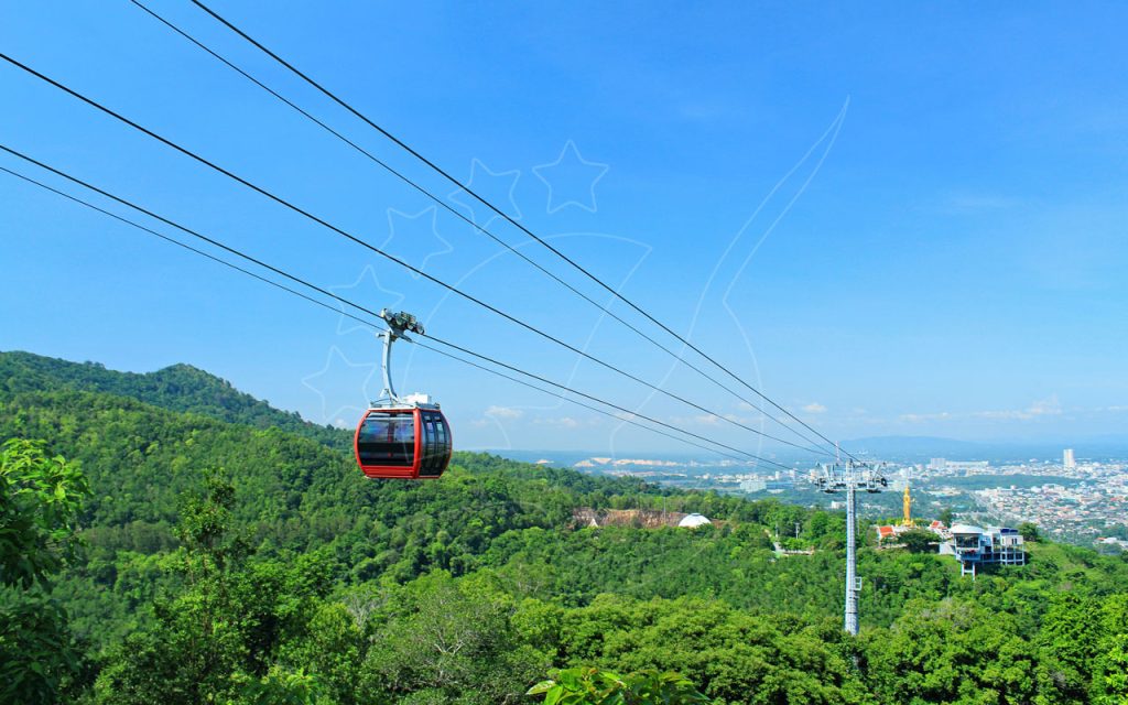 Red cable car gondola over lush green hills with a coastal city and golden statue in the distance