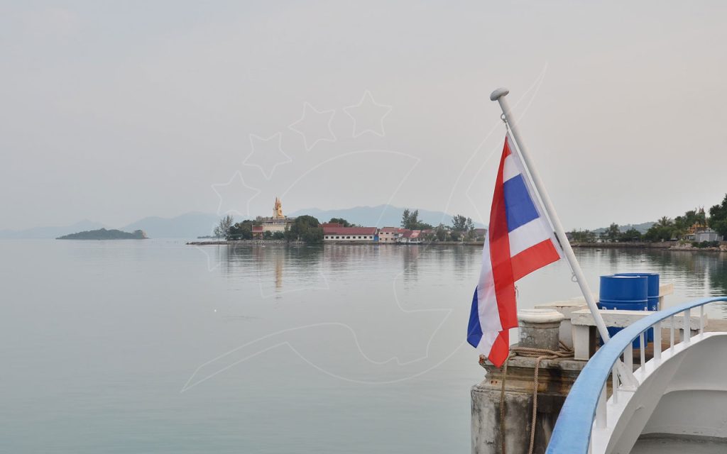 Thai flag on a pier railing overlooking calm hazy sea with distant temple and golden Buddha on a headland