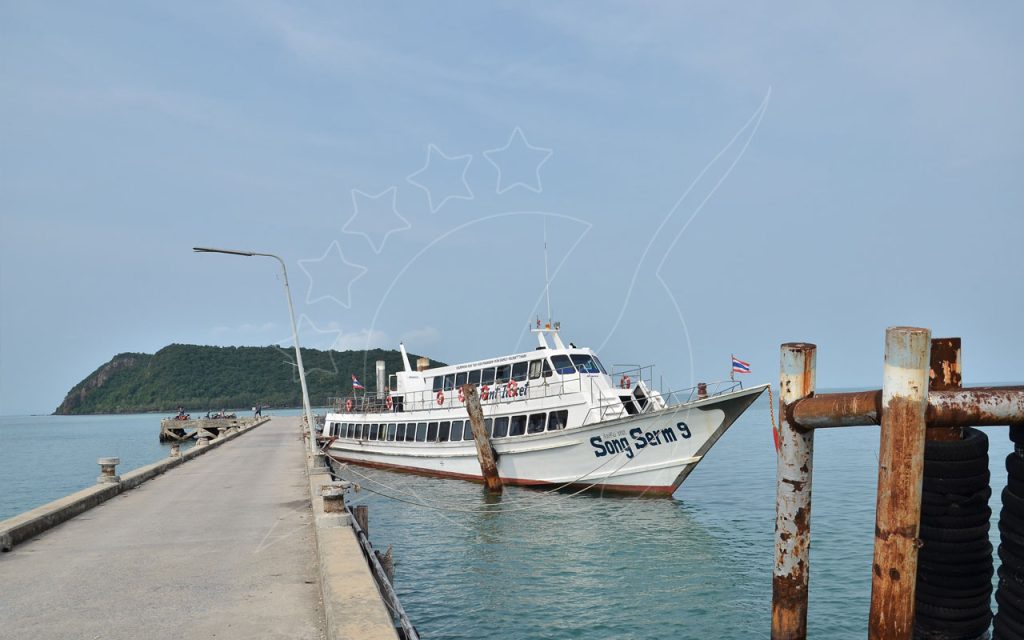Songserm ferry moored at a long concrete pier under hazy blue sky, green island headland in the distance