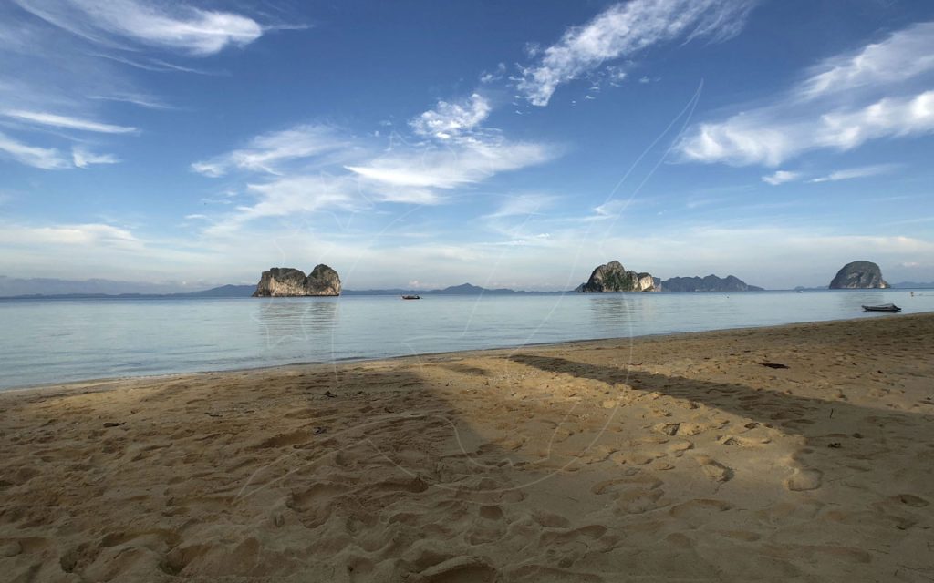 Empty sandy beach at low tide with calm sea and limestone karst islands under a blue sky with wispy clouds