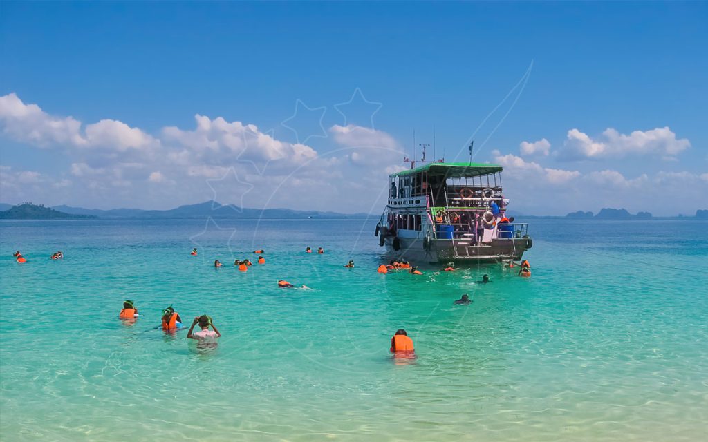 Snorkellers in orange life jackets float in clear turquoise shallows beside a wooden tour boat off a tropical island