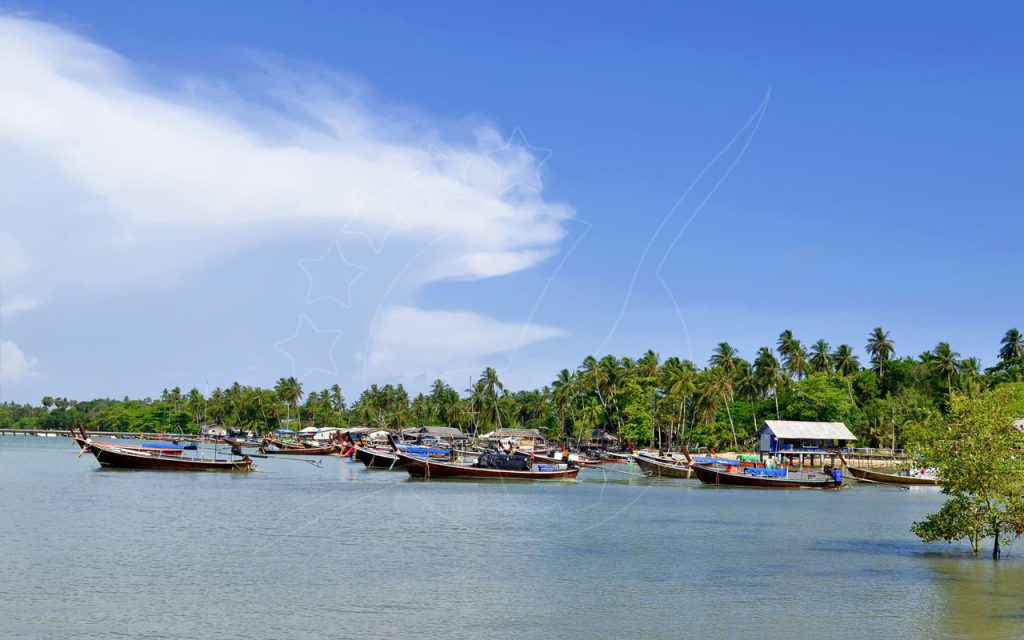 Wooden longtail boats moored on calm water beside a palm-fringed fishing village under a blue sky