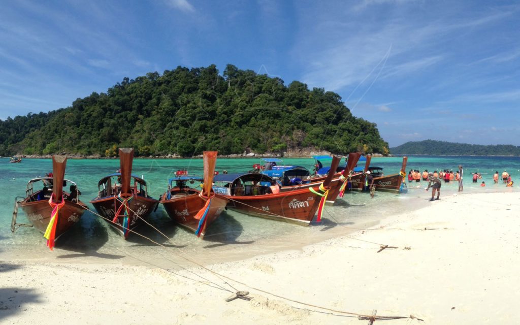 Wooden longtail boats moored on a white sand beach with turquoise water and a jungle-covered island behind