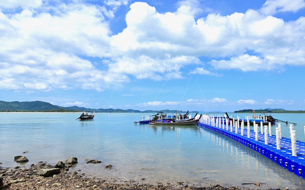 Blue floating plastic pontoon pier with moored longtail boats on calm turquoise water under bright cumulus clouds