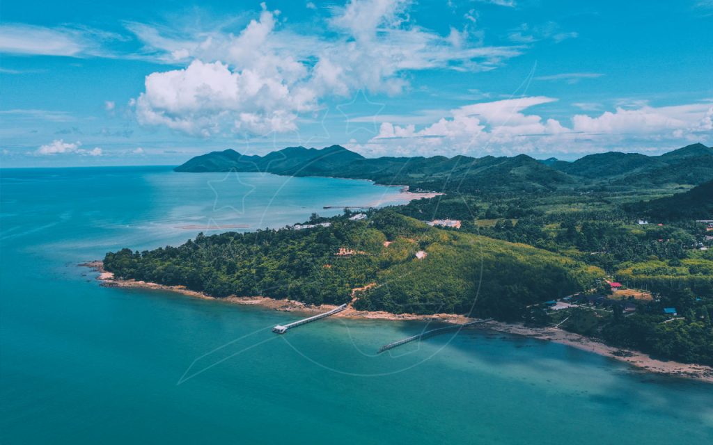 Aerial view of a tropical green headland with wooden piers, turquoise sea and forested hills under blue sky