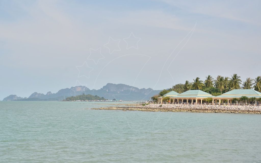 Green-roofed pavilions and palm trees on a rocky coastal breakwater with limestone karst hills across hazy calm sea