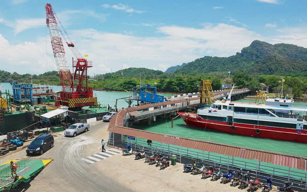 Ferry pier in southern Thailand with docked passenger boat, parked motorbikes, red crane and limestone hills