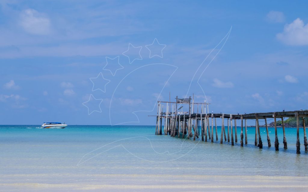 Weathered wooden pier stretching into turquoise shallows with a speedboat anchored offshore under a blue sky