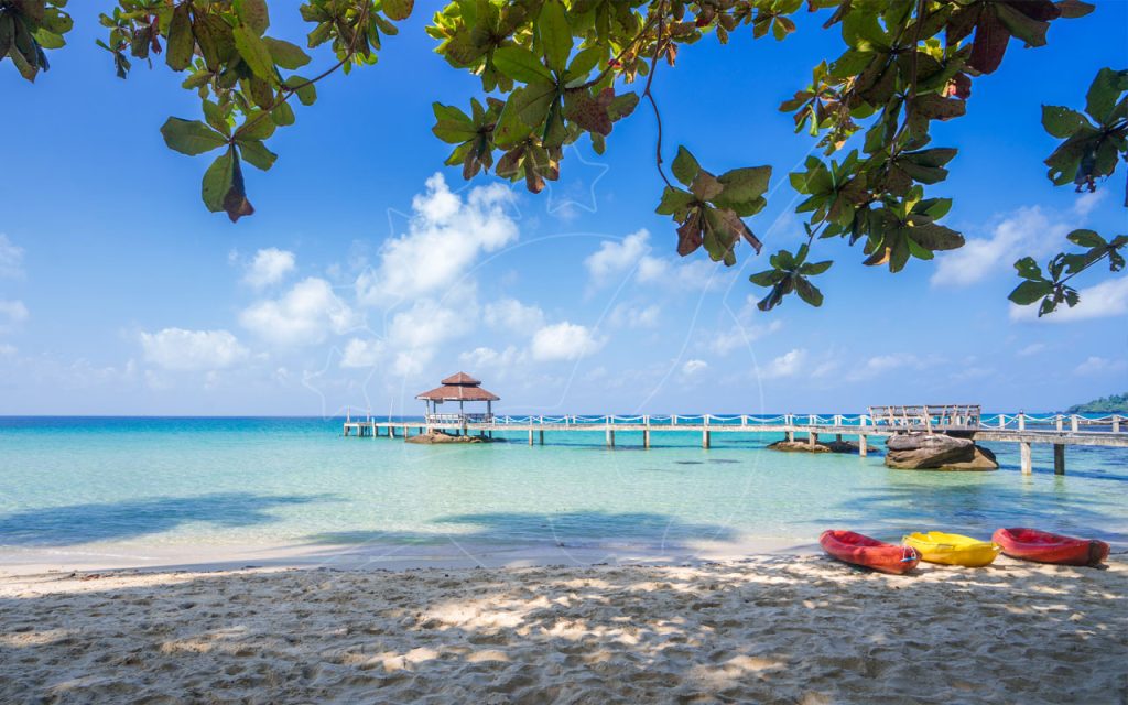 Wooden pier with red-roofed gazebo over turquoise water, three kayaks on white sand beach framed by leaves