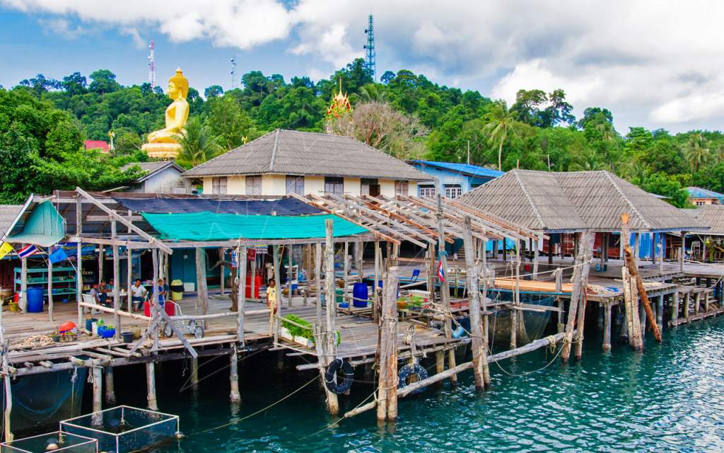 Stilted wooden fishing village over turquoise water with hilltop golden Buddha and jungle behind, Thailand