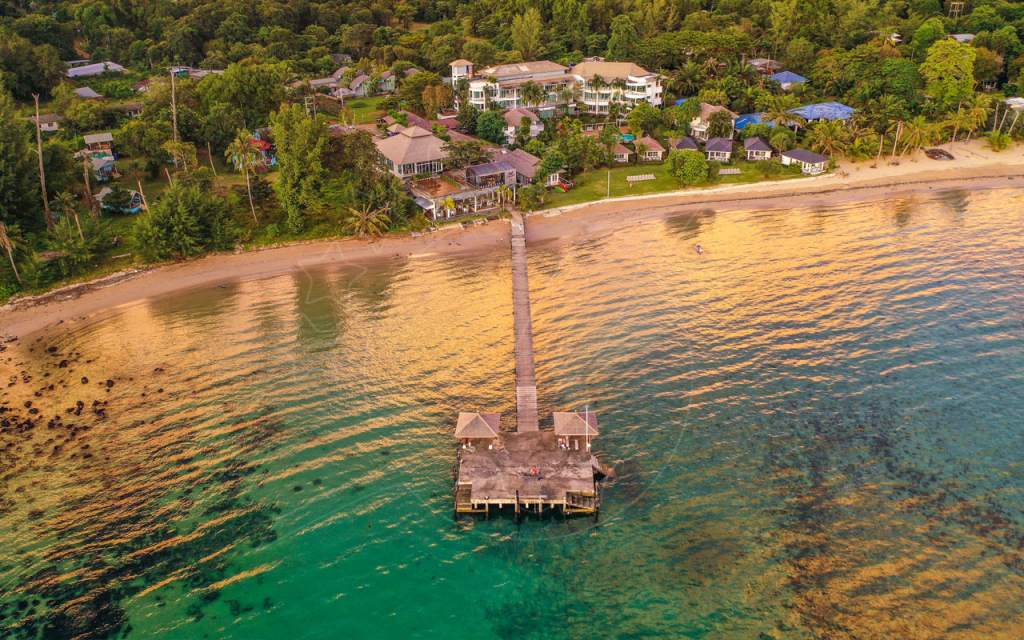 Aerial view of a long wooden pier with two pavilions reaching into turquoise water beside a beachfront resort