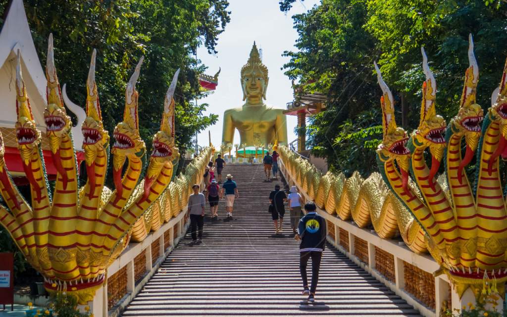 Golden naga serpent balustrades line a temple staircase leading to a large seated golden Buddha statue, Thailand