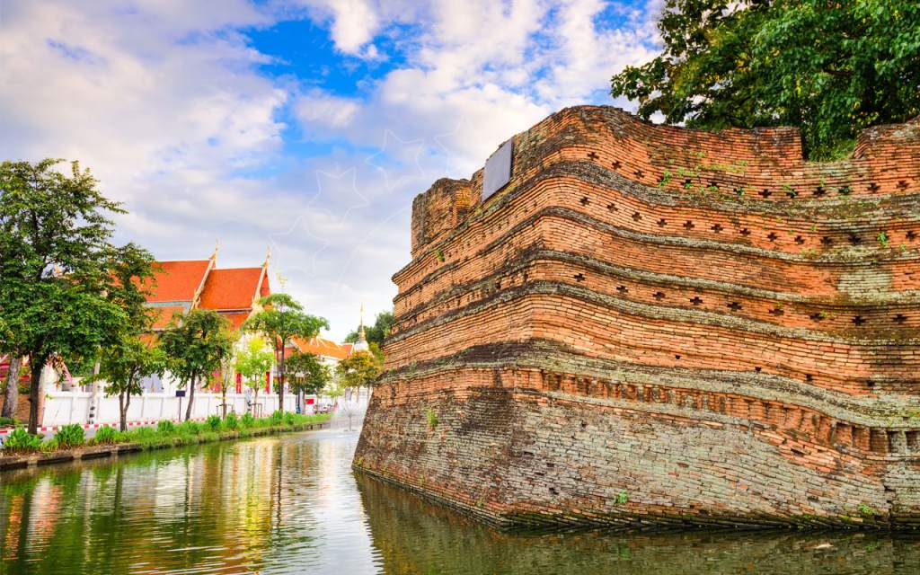 Weathered red brick corner of Chiang Mai's old city wall beside a green moat, with a Thai temple roof and trees behind