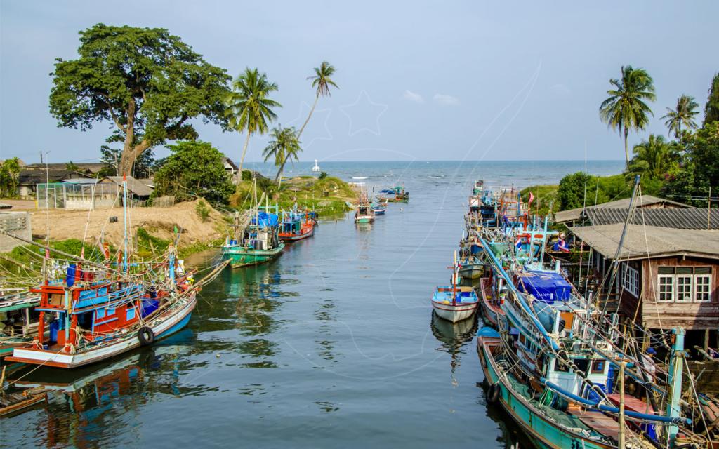 Colorful wooden fishing boats moored along a canal leading to the sea, with palm trees and stilt houses