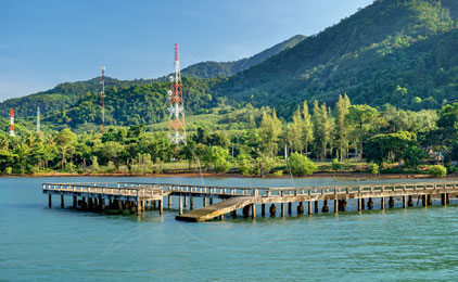 Wooden pier with yellow boat extending into turquoise water, forested mountains and red communication tower in background