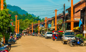 Thai town main street lined with orange-pillared shopfronts, parked motorbikes and pickup trucks, green hills behind