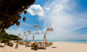 Beachfront restaurant tables under thatched parasols on white sand beside turquoise sea and blue sky