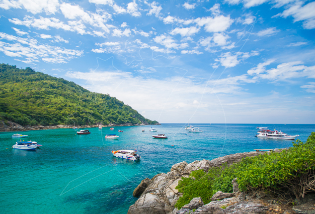 Speedboats and a dive boat anchored in a turquoise tropical bay beside a forested island and rocky headland