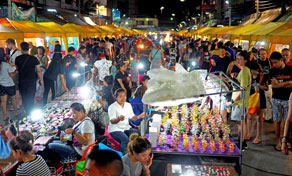 Crowded night market at dusk with lit stalls, vendors and shoppers browsing souvenirs and trinkets under canopies
