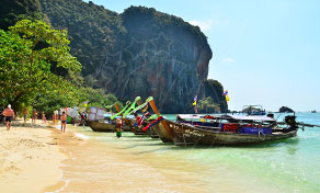Longtail boats moored on a white sand beach beneath a limestone karst cliff in Thailand