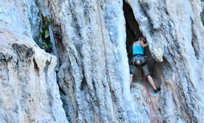 Climber in blue tank top ascending a grey limestone cliff face with rope beside a dark cave opening